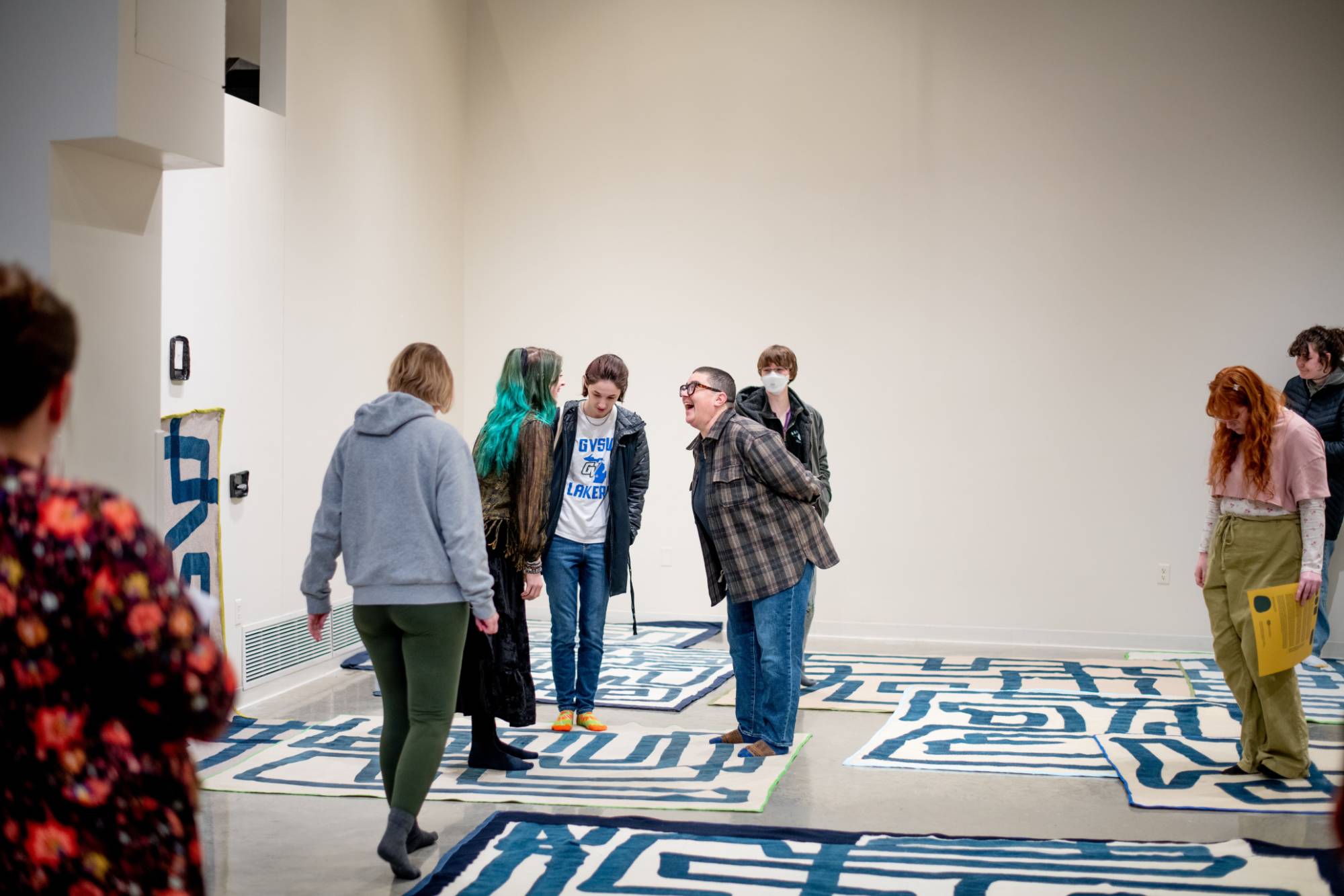 Visitors to the Padnos Gallery standing on an art installation by Emmy Bright, made up of wool rugs drawn on by a printmaking process to look like lines.